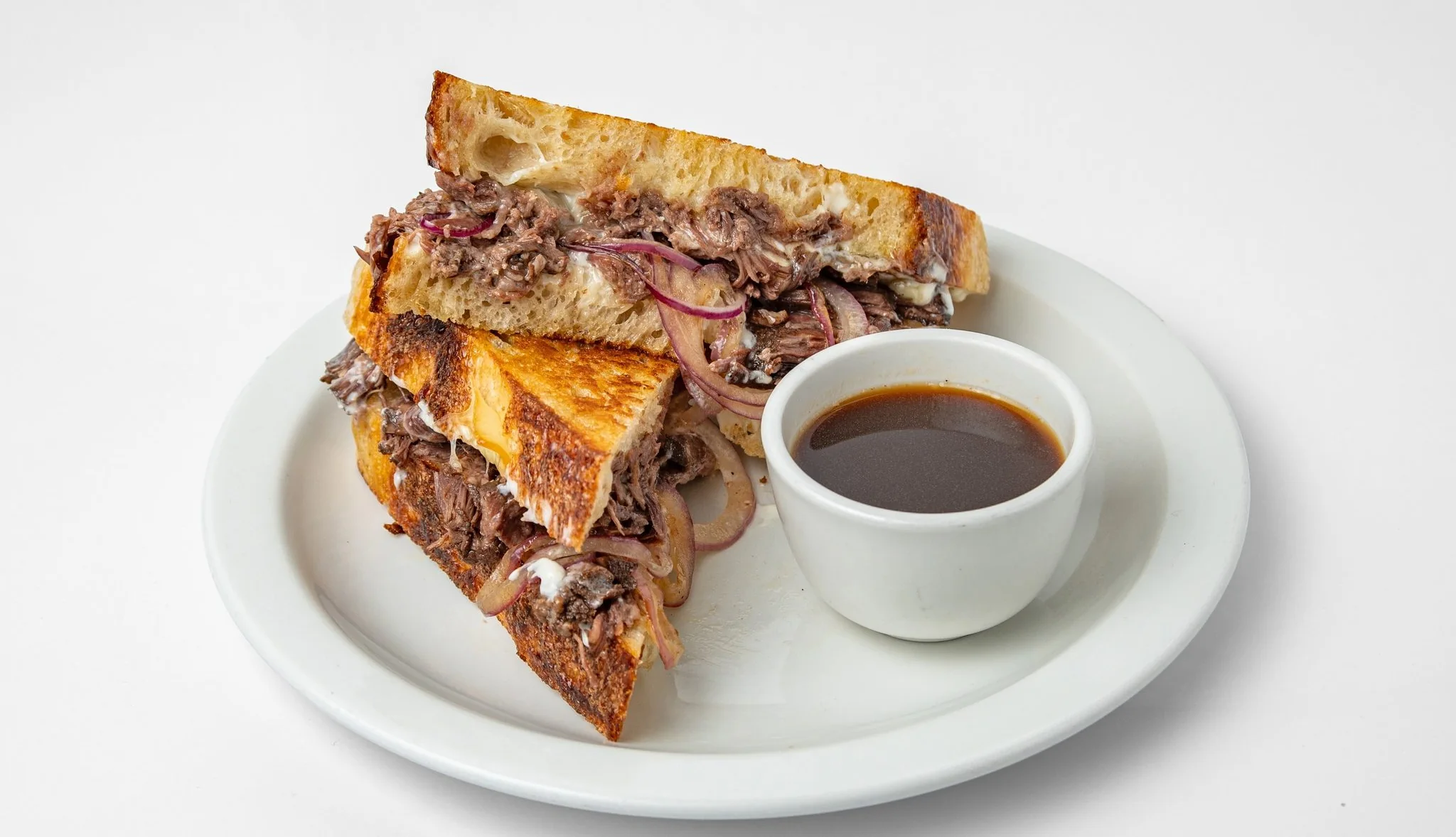 Bistek beef dip on toasted sourdough with a cup of jus on a white plate